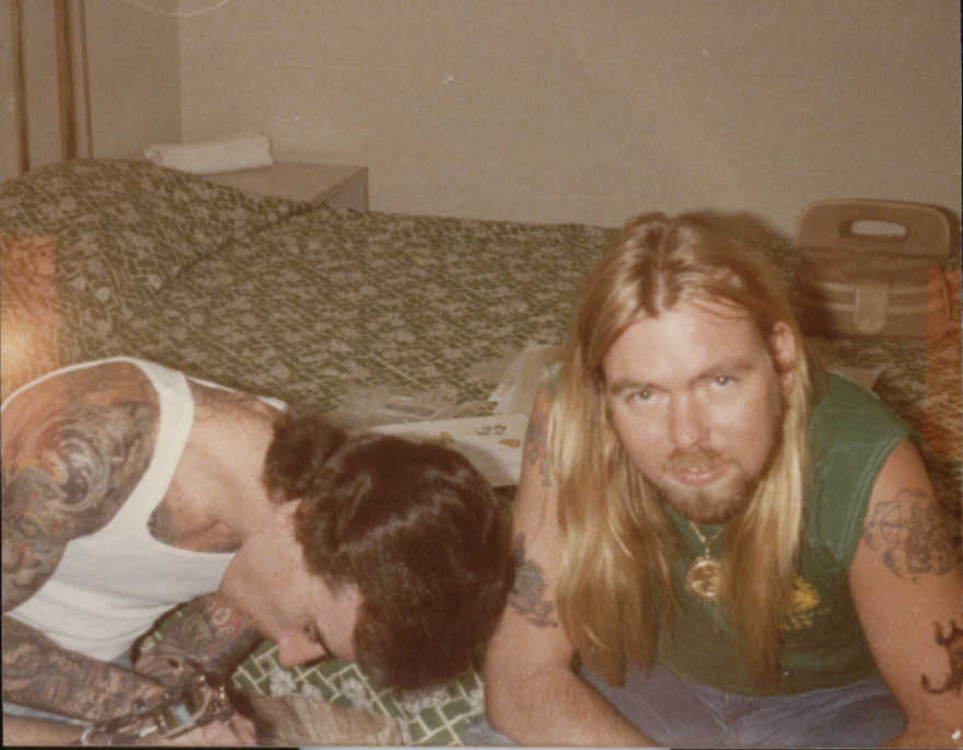 Neil Grant tattooing Jim Essery (harp player) for the Enlightened Rogues Tour, while Gregg looks on after just getting tattooed. 6-25-79. 